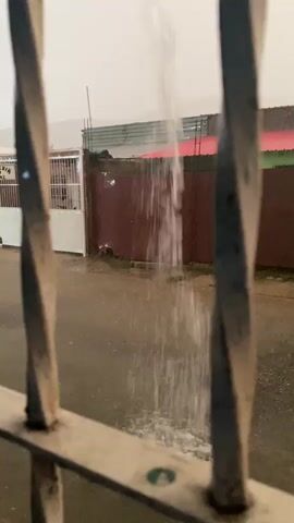 A stream of water falls from an unseen source above, splashing onto a wet asphalt surface. The water is falling in a narrow column, breaking into a spray as it hits the ground. In the background, a red and green metal fence runs along a brown wall. To the left of the fence is a white gate with a white building behind it. The scene appears to be outdoors, possibly during a rainstorm, with the ground being wet and reflective. The foreground is dominated by the blurred image of what appears to be a metal grate or bars, obscuring the lower portion of the view. The overall lighting is dim, suggesting an overcast day or evening.