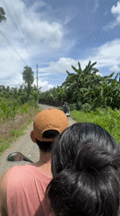 Moped riders travel dirt road through banana trees in Carmen