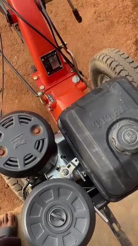 A video shows a red and black walk-behind tractor with two wheels. The tractor has a black gas tank with the word "TITAN" visible on it. There are two round black air filters on the left side of the tractor. The tractor appears to be outdoors on a dirt surface. Some water bottles and a bag are attached to the frame of the tractor. The footage is shot from a high angle looking down at the tractor.