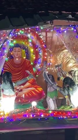 The video shows a colorful display, likely religious, at night. A large image of a seated Buddha, wearing a red robe, is in the center, framed by a colorful swirling background and strings of colorful lights. In front of the Buddha, three figures are depicted. They are dark-skinned, and appear to be performing a dance or ritual, wielding props like axes or swords. The scene is illuminated by the colorful lights, and a roof structure is partially visible in the background, suggesting the display is located outdoors, possibly on a building or platform.