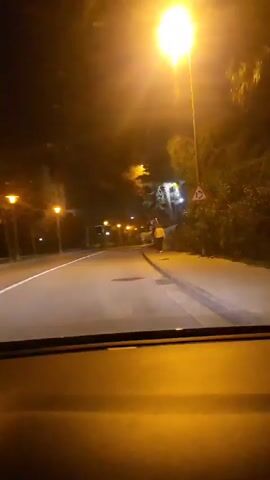 A video shows a car driving on a street at night. Streetlights illuminate the road. A person in a yellow jacket walks on the sidewalk. The road has a white line marking the edge of the lane. There are trees and a building visible along the side of the road. The video is shot from the perspective of the driver.