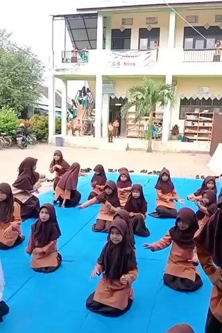 A group of young girls in brown and black hijabs and dresses are kneeling on a blue mat outdoors. They are performing some kind of coordinated movement, possibly a dance or exercise routine. An adult woman, also wearing a hijab and a brown dress, is standing in front of them, seemingly instructing them. In the background, there is a multi-story building with columns and windows, suggesting it might be a school or community center. Other children and adults can be seen in the background. The ground is paved, and there are some bicycles parked to the left. The overall scene appears to be an outdoor educational or recreational activity.