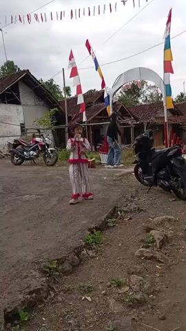 A child in a pink and white patterned pajama set stands in a dirt road, holding a flute and looking to the right. The child has dark hair pulled back. Red and white Indonesian flags are visible on poles behind the child. An archway decorated with Indonesian flags is further back. A black motorcycle is parked to the right of the child, and a red and white motorcycle is parked to the left. Several houses with tile roofs are in the background. Two people are standing near the archway. The ground is dirt and rocks with some green vegetation. The sky is overcast.