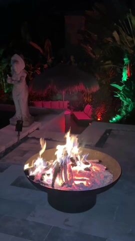 The video shows a fire burning in a round, dark-colored fire pit, with flames licking up from the logs. The fire pit sits on a stone-tiled patio, and behind it is a statue lit with red light, a small tiki-style umbrella, and some greenery illuminated with red and green lights. The overall scene is dimly lit, suggesting it is nighttime.