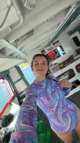 The video shows a young woman standing indoors, facing the camera. She has curly brown hair and is smiling. She is wearing a long-sleeved swimsuit with a purple and blue swirled pattern. The background appears to be the interior of a boat or a similar structure, with wooden beams on the ceiling and windows showing the ocean and some land in the distance. There are some items visible in the background, including shelves with various objects and a sign that says "NO SMOKING." The woman is holding her hand to her swimsuit.