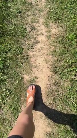 This video captures a first-person perspective of someone walking along a narrow, sandy path through a grassy, rural landscape. The sun is shining brightly, suggesting it is daytime, and the weather appears to be clear with a few clouds visible in the distance. The path is surrounded by lush green grass and some scattered wildflowers. In the background, a line of trees can be seen against the bright sky. The person is wearing brown sandals. The scene evokes a sense of calm and natural beauty, possibly indicating a leisurely walk in the countryside. The location context provided is Шолохово, Russia. There is no visible text in the video.