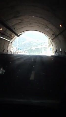 The video begins from inside a vehicle as it exits a tunnel. The perspective is from the driver's seat, looking forward through the windshield. The tunnel's curved, concrete ceiling and walls are visible at the beginning of the clip, along with lighting fixtures on the left side. As the vehicle emerges, a multi-lane highway comes into view under a bright, clear sky. The highway is paved with asphalt and marked with white lane lines. Road signs are visible on both sides of the highway, including blue directional signs with white symbols and text. The landscape outside the tunnel consists of rolling hills covered with green vegetation and sparse trees, indicating a rural or semi-rural setting. The sun is shining, casting shadows on the road. There are no other vehicles or people clearly visible in the immediate vicinity as the car exits the tunnel.