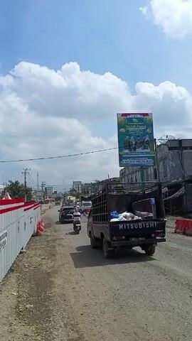 The video shows a view of a street in what appears to be a small town. The road is unpaved and partially under construction, with barriers lining one side. Several vehicles, including a black Mitsubishi pickup truck and motorbikes, are traveling on the road. There are buildings on either side of the street, some of which look like shops or businesses.  A large billboard is visible, displaying a photograph of several people with a message in Indonesian, likely related to a religious holiday. The sky is blue with fluffy white clouds. The overall impression is a busy, but somewhat underdeveloped area under a clear sky.