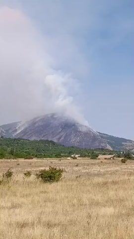 This video shows a mountain with a significant amount of smoke rising from its peak. The mountain slope appears to be scorched in several areas, indicating a recent or ongoing fire. The foreground consists of a dry, grassy field with scattered bushes. In the distance, there are trees and some buildings, suggesting a rural or semi-rural environment. The sky is partly cloudy.
