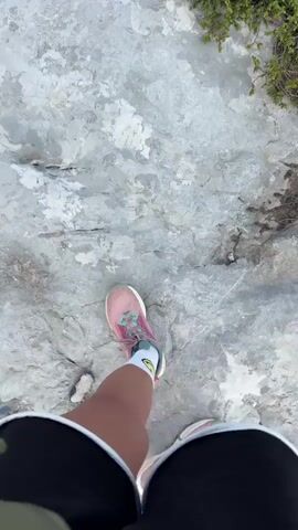 This video shows a POV perspective of someone walking on a rocky, mountainous terrain. The person is wearing pink and white athletic shoes and black shorts. The path is uneven and covered in large, grey rocks. In the background, there are lush green trees and distant mountain ranges under a cloudy sky. The camera pans up to reveal a vast landscape of mountains and valleys.