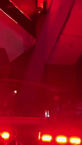 The video begins with a close-up view of a stage bathed in red light. A large, angular structure, possibly part of the stage design or a building, is visible in the background. As the video progresses, beams of white light begin to sweep across the scene, creating a dynamic visual effect. Trees are visible on the left and right sides of the frame, illuminated by the red lights. A crowd of people can be seen on the stage, some appearing to be DJs or performers interacting with equipment. The atmosphere suggests a concert or music event with a strong emphasis on lighting and visual effects. The video ends with the scene entirely engulfed in darkness.