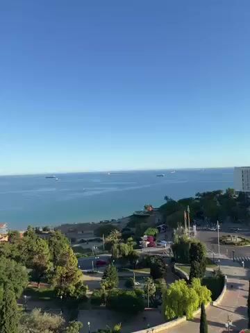 The video presents a high-angle panoramic view of Tarragona, Spain, under a clear, bright blue sky. It opens with a wide shot of the Mediterranean Sea, where several large ships or vessels are visible in the distance on the calm water. The coastline features a beach, green spaces with trees, and a large white building on the right.

The camera then slowly pans right, revealing more of the city. A prominent multi-story white building, possibly a hotel, overlooks the sea. Below it, a well-maintained urban area includes a roundabout with traffic, surrounded by trees and landscaped gardens. Ancient stone walls or ruins, suggesting the city's historical significance, are also visible, contrasting with the more modern structures.

As the pan continues, a sprawling cityscape unfolds, featuring numerous residential and commercial buildings with varied architectural styles and roof colors. A distinct large pink building stands out in the mid-ground. The sun is clearly visible in the sky towards the end of the video, confirming it is daytime with excellent weather conditions. There is no discernible text visible in the video.
