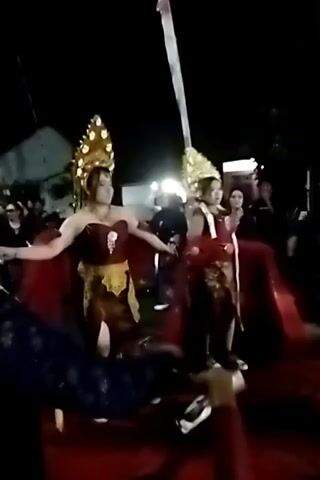 The video shows a nighttime outdoor event, likely a celebration or performance. In the foreground, two individuals are dressed in elaborate traditional attire, including ornate golden headpieces and red capes. They appear to be dancing or posing. The woman on the left is wearing a maroon top and a yellow and brown skirt with a split. The person on the right is wearing a maroon top and dark pants. There are other people in the background, some of whom are also dressed in traditional clothing. The ground is covered in a red carpet. A tall, white flag on a pole is visible in the background. The lighting suggests artificial illumination, with some sources appearing to be spotlights.