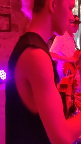 This video captures a music performance in a dimly lit room, illuminated by pink and purple lights. In the foreground, a person wearing a black sleeveless shirt is playing a violin. To their right, another person is playing a bass guitar. Behind them, a person in a white polo shirt and a baseball cap is also playing the bass guitar. Further back, a man in a black hoodie and gray pants stands observing. The background features exposed brick walls adorned with posters and artwork. Several electronic music devices, including keyboards and mixing boards, are set up on tables in the middle of the room. The video pans to reveal other individuals seated and standing, observing the performance. A woman in a blue jumpsuit is seen singing into a microphone. The overall atmosphere suggests a live music event or practice session with a band.