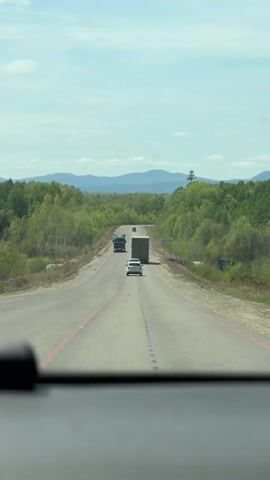 The view from inside a car looking through the windshield shows a straight, paved road with cracked gray asphalt flanked by green deciduous trees. In the distance are blurry mountain ranges beneath a blue sky with patchy white clouds. A large dark truck and a white car are moving away from the camera, with the truck closest and a flatbed truck further down the road. The road has red lines on the left and right edges. A signpost with illegible lettering is on the right side of the road. A portion of the dashboard can be seen in the lower part of the frame.