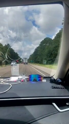 A dashboard camera records a car's journey on a multi-lane highway under a partly cloudy sky. The road is lined with green trees on both sides. A tractor-trailer and a white sedan are visible ahead. A green sign reads "Weigh Station 1 Mile". The car's dashboard holds sunglasses, a charging cable, a sparkly object, and some papers.