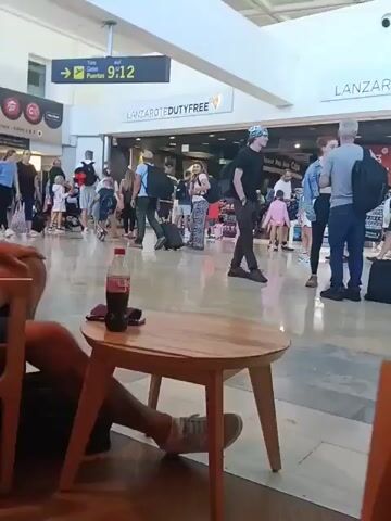This video captures a busy scene inside an airport terminal. Numerous people of various ages, including adults and children, are seen walking across a brightly lit, tiled floor. Many individuals are carrying or pulling luggage, such as suitcases, backpacks, and duffel bags, indicating ongoing travel activity.

In the immediate foreground, two individuals are partially visible, seated in chairs, with a small, light-colored wooden table between them. On the table rests a bottle of Coca-Cola and a pair of sunglasses.

Prominent signage in the background provides key location details: a large sign reads "LANZAROTE DUTY FREE," clearly identifying the airport as Lanzarote Airport. Another overhead sign displays "Gates / Puertas 9-12" with an arrow, directing passengers to boarding gates. The overall lighting is bright and even, consistent with an indoor public space like an airport. The activity primarily involves people moving about, likely waiting for flights or having just arrived. The scene appears to be active and somewhat crowded, but no specific events or strong emotions are discernible among the travelers.