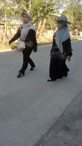 A video taken outdoors shows a group of people gathered near a street. In the foreground, a person is walking away from the camera. The person is wearing a black jacket, black pants, a grey headscarf, and a wide-brimmed hat. They are holding a white object in their right hand. The ground is paved with concrete. Behind this person, other individuals are visible, some sitting and some standing. Several motorcycles are parked along the side of the street. Trees and foliage can be seen in the background. The lighting suggests it is daytime. The video appears to be shot from a slightly low angle and is oriented vertically.