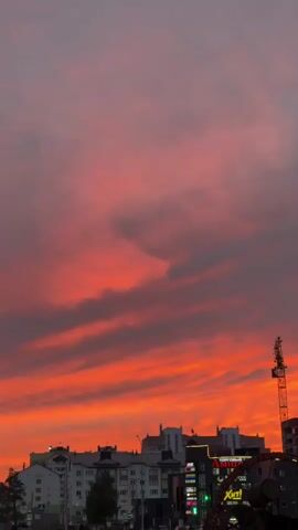 The video shows a city skyline during a vibrant sunset. The sky is filled with dramatic red and orange clouds, with streaks of darker gray clouds interspersed. In the foreground, silhouetted buildings are visible, including apartment buildings and what appears to be a commercial building with illuminated signs. A construction crane stands tall on the right side of the frame. The overall mood is serene and picturesque, capturing the beauty of nature contrasting with the urban environment.