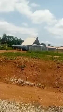 The video begins with a shot of a dusty, red dirt road with some buildings and large metal tanks in the background. The camera pans to the right, revealing more buildings and some vegetation. The scene is an outdoor environment with a partly cloudy sky. The camera continues to pan, showing a long, beige wall with a barbed wire fence on top. Two women are walking along the side of the road. The first woman is wearing a white t-shirt and a patterned skirt, and the second woman is partially visible and is also wearing a patterned skirt. The camera continues to pan, showing more of the wall and the road. The video appears to be taken from a moving vehicle.