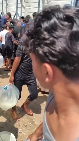 A group of people, including children, are gathered in a crowded outdoor area, possibly a street or an open space next to a building. They are holding buckets and plastic containers of various sizes, some of which are empty, while others appear to be filled or in the process of being filled with liquid. Many individuals are wearing casual clothing, and some are barefoot. The atmosphere seems to be one of urgency and shared effort, with people passing containers and interacting with each other. The background consists of a white, textured wall with some metal railings or structures. The ground is rough and appears to be dirt or gravel. The lighting suggests it is daytime, with sunlight casting shadows. Some people are looking down, focused on filling or carrying their containers, while others are looking towards a source of the liquid or at each other.