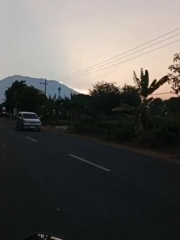 The video shows a view from a motorcycle traveling on an asphalt road. The sun is low in the sky, casting a warm glow. In the distance, a mountain is visible, partially obscured by clouds. Power lines stretch across the sky, and trees and vegetation line the sides of the road. A silver car passes by in the opposite direction, and a red motorcycle with a rider wearing a dark jacket and headscarf is also seen. The overall atmosphere is serene, suggesting a quiet rural setting at dusk or dawn.