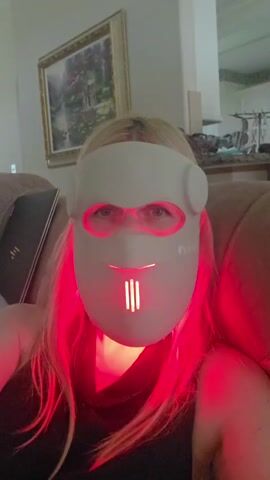 A woman is sitting on a couch, wearing a white LED facial mask that emits a red light. The mask covers her entire face, with openings for her eyes and mouth. The red light from the mask illuminates her face and hair, casting a glow around her. She has blonde hair that falls around her shoulders. The background appears to be a home interior, with a painting on the wall and shelves with items in the distance. The overall impression is of a personal care routine taking place in a casual setting. There is no text visible in the media file.