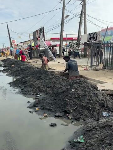 The video shows a street scene in Lagos, Nigeria, with a focus on a drainage ditch that has been cleared of a large pile of dark, muddy debris. Several people are visible, including workers engaged in the cleanup and onlookers.

In the foreground, two young men are actively working. One, wearing a blue t-shirt and red shorts, is using a shovel to move the mud. He appears to be looking towards the camera. Another young man, in a plaid shirt and tan trousers, is standing near the edge of the ditch, holding a shovel. He looks directly at the camera and smiles, then turns and gestures with his head towards something off-camera. Further back, another person in a red shirt is also seen near the ditch.

The background reveals a busy street with various buildings and businesses. Signs are visible, including one that reads "HIPCO." Clothes are hung out to dry on a line. There are also various people in the background, some standing around and others appearing to be customers or vendors. The general atmosphere suggests a bustling urban environment.

The time of day appears to be daytime, with bright but possibly hazy sunlight, judging by the lighting and the presence of shadows. The weather seems to be warm and possibly humid, typical of Lagos. The scene depicts a common urban maintenance activity, likely related to clearing clogged drainage systems to prevent flooding. The smiles of the workers suggest a sense of camaraderie or a lighthearted moment despite the difficult nature of the work.