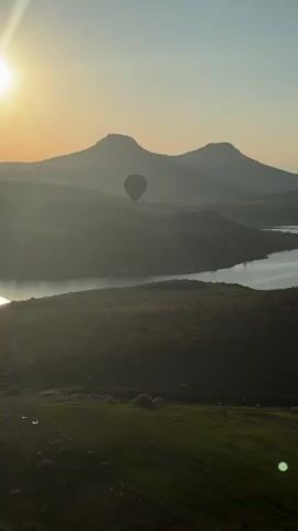 The video captures a serene sunrise or early morning scene in Yeşilhisar, Turkey, from what appears to be a hot air balloon or a high vantage point. The sky transitions from a soft, warm glow on the left to a pale blue overhead, casting gentle light across the landscape. Rolling hills and mountains dominate the background, partially obscured by a light atmospheric haze, while a winding body of water, possibly a river or reservoir, reflects the sky.

In the foreground and mid-distance, agricultural fields with green crops and patches of bare earth stretch across the terrain. Throughout the video, several hot air balloons are visible, adding to the picturesque scene. Initially, a single dark-colored balloon is seen floating over the water and distant mountains. As the camera slowly pans right, other balloons emerge into view, including a prominent yellow one and another dark-striped balloon, flying gracefully over the varied landscape and near distinct rocky formations. The distinctive whooshing sound of a hot air balloon burner is frequently heard, underscoring the aerial perspective of the footage. The overall impression is one of peaceful flight over a beautiful and expansive natural environment.