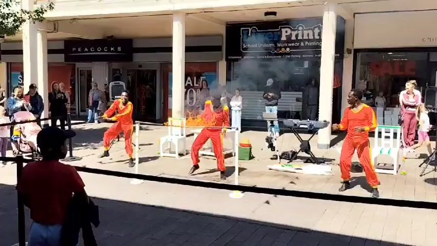 The video shows an outdoor performance on a paved street, likely in a shopping area with storefronts visible in the background. Three individuals dressed in bright red outfits are performing a stunt. The central performer appears to be engulfed in flames, with smoke rising from their upper body. This performer is positioned between two other individuals who are also in red outfits and are participating in the performance, possibly holding something or reacting to the flames. Two white tables are placed in front of the central performer. In the background, several onlookers are watching the performance, some clapping. Storefronts labeled "PEACOCKS" and "Live Print" are visible. The lighting suggests it is daytime with clear weather.