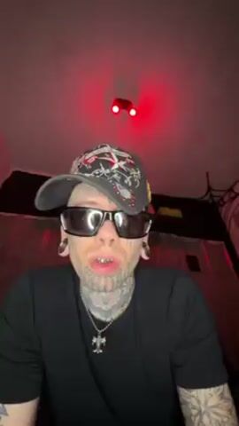 A person with extensive face tattoos, wearing sunglasses and a baseball cap, is seen from a low angle. They are wearing a black t-shirt and a silver necklace with a cross. The person opens their mouth to reveal metallic, perhaps gold, teeth. Behind them is a dimly lit area with red lighting, and some objects are visible, including what appears to be a corrugated metal surface, a spiderweb decoration, and a large white object. The camera is relatively still.
