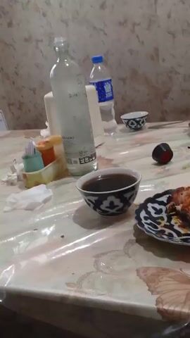 The video shows a table set with food and drinks. The table is covered with a clear plastic tablecloth with a marble-like pattern. There are several plates with food, including what appears to be fried food and salads. There are also small bowls with a traditional blue and white pattern, some containing a dark liquid, possibly tea. A tall glass bottle of water and a smaller plastic bottle are visible. A green plastic basket containing bread is also on the table, along with a cell phone. The background appears to be a simple wall. The video is shaky, indicating it was likely taken by hand. There are no people clearly visible in the frame.