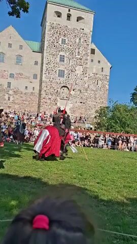 A reenactment of a medieval jousting tournament is underway. Two knights on horseback, adorned in armor and decorative caparisons, are charging towards each other. One knight is on a black horse with a red blanket, and the other is on a white horse. They are on a grassy field with a large stone castle in the background. Spectators are gathered behind a red barrier, watching the event. The sky is clear and blue.