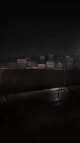 The video, shot at night, depicts a scene dominated by stacked shipping containers. The containers, arranged in multiple rows, appear in various colors, including green, yellow, and red. A dark barrier, possibly a wall or embankment, partially obscures the lower portion of the containers. The foreground is dark, with a road or path visible, glistening from rain or wet conditions. Sparse vegetation or branches are seen along the edge of the path. Overall, the scene is dimly lit, adding to the somber atmosphere.