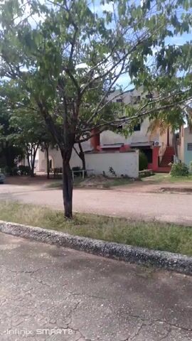 The video shows a street with apartment buildings on either side. The buildings are two or three stories tall and appear to be residential.  The street is paved but cracked, with a stone or concrete curb along the side. There is grass and some small trees alongside the road. The sky is blue with scattered clouds. A motorbike passes in the distance. The video is taken from a moving vehicle.