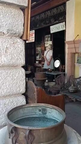 The video shows the storefront of an antique shop. Two elderly people enter the shop. The exterior is decorated with various antique items, including a large copper pot filled with water, stone blocks, a wooden bench, a cast iron bench, and a painting of a cityscape above a mantle. The sign above the shop reads "ANTIQUES OLD-NEW". Another sign to the left says "VISITATE PALAZZO MUSEO".
