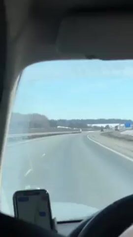 The video shows a view from inside a car driving on a highway. The car is moving forward. The sky is blue and there are patches of snow on the side of the road. There are some trees in the distance. A smartphone is attached to the car dashboard, displaying a map. The steering wheel is visible.