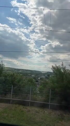 This is a video clip captured from a moving vehicle, likely a train, as evidenced by the blur of the landscape and the presence of power lines in the foreground. The scene depicts a rural or semi-rural landscape under a dynamic sky.

**Subjects and Setting:** The primary subjects are the natural landscape and the sky. We see lush green trees, forming a dense canopy that partially obscures a village or town in the distance. The village appears to consist of houses with red and white roofs, nestled amongst the trees. Beyond the village, rolling hills are visible, some appearing to be scarred by quarrying or construction. In the very foreground, a metal fence and some dry grass are visible, suggesting the edge of a field or embankment. There are no people visible in the frame.

**Activity or Event:** The video captures a moment in time as the viewer is transported through this landscape. The sense of movement is key, with the scenery subtly shifting and the power lines remaining relatively static overhead.

**Notable Details:**
*   **Time of Day:** The bright, varied lighting on the clouds suggests it is daytime, likely late morning or early afternoon.
*   **Weather:** The sky is filled with large, fluffy cumulus clouds interspersed with patches of blue sky. This indicates partly cloudy weather, possibly with a pleasant temperature.
*   **Location Cues:** The mention of "Liski, Russia" as the location context provides a specific geographical setting. The architectural style of the houses (red and white roofs) and the general topography are consistent with rural Russian landscapes. The presence of utility poles and wires further supports this.
*   **Emotions:** The scene evokes a sense of calm and transit, typical of travel. The vastness of the sky and the greenery suggest a peaceful, natural environment.

**Visible Text:** There is no visible text within the video frame itself. The timestamps at the beginning of the clip are part of the metadata, not the content of the recording.