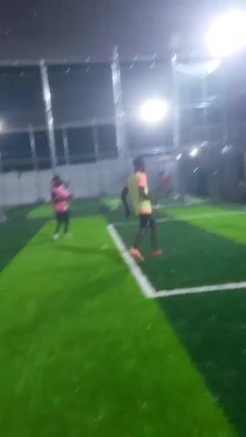 A medium shot captures a soccer game in an indoor facility at night. Several young men are visible on a green artificial turf field with white lines. Bright overhead lights illuminate the area. A tall metal fence encloses the playing space. In the foreground, one player in a pink and light blue jersey and black shorts walks away from the camera. In the middle ground, two players, one in a pink jersey and black shorts, and another in a yellow jersey and blue shorts, are actively playing with a white soccer ball. Other players in various colored jerseys and shorts are visible in the background, also engaged in the game.