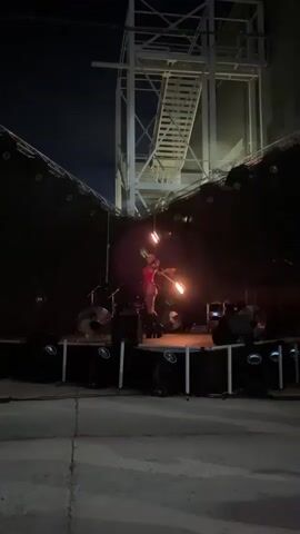 A person dressed in a red costume with black accents and tall black boots stands on a stage performing with fire. The person is holding a flaming prop in their right hand and appears to be swinging it. The stage is dark with various lights and what look like drum set components in the background. A large metal structure with stairs is visible behind the stage. The overall environment appears to be an outdoor nighttime setting.