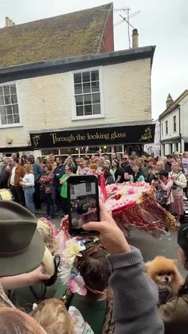 A crowd of people lines both sides of a street as a parade float approaches. The float features a smiling young woman with pink hair and a sparkly outfit, seated on a round platform decorated with flowers and streamers. Many people in the crowd are holding up their phones to record the float as it passes.  A small, fluffy dog is visible in the arms of a woman in the foreground.  The building behind the crowd on one side of the street has a sign that reads "Through the looking glass." The day is overcast.