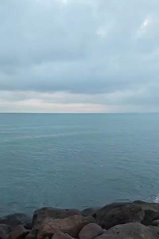 The video shows a wide shot of the ocean on a cloudy day. In the foreground, there is a rocky shore line. The water is a murky blue-green color. The sky is covered in grey clouds, with a hint of light peeking through in some areas. The scene is calm and still, with only slight ripples visible on the water's surface.