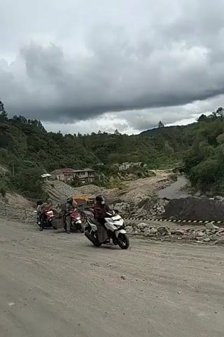 The video shows a rural scene in Jamurujung, Indonesia, likely during the daytime under a cloudy sky. The foreground is dominated by a dirt road, and a person on a white scooter is riding towards the right of the frame. Further back, three other scooters are parked or moving slowly on the same road. The scooter closest to the camera has a rider wearing a helmet and a dark jacket. The second scooter has two people on it, one wearing a helmet and the other a hijab. The third scooter, partially obscured, also appears to have two riders.

The middle ground of the scene reveals a construction or excavation site. A river flows through a rugged, rocky area where piles of dirt and debris are visible. Construction equipment, including a yellow excavator, is present near the river. Yellow and black hazard tape marks off sections of the site, suggesting ongoing work or a hazardous area. In the background, lush green hills rise, dotted with trees and some buildings, possibly homes. The overall atmosphere appears somewhat overcast, with dark clouds suggesting the possibility of rain. The activity depicted is primarily travel and possibly observation of the construction work. There is no visible text in the video.