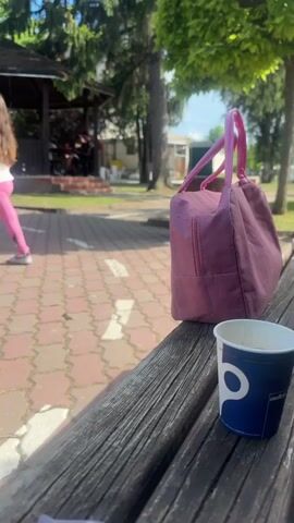 The video depicts a young girl in a white t-shirt and pink leggings walking on a brick-paved pathway in a park. She is wearing roller skates and is moving from left to right in the frame. In the foreground, a wooden bench holds a purple lunch bag and a blue and white paper cup. Behind the girl is a gazebo with people sitting inside. Trees are visible in the background, and the overall scene suggests a sunny day in a public park or recreational area.
