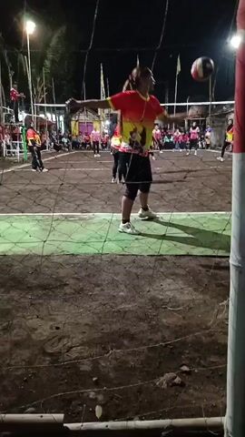 A nighttime volleyball game is being played outdoors on a dirt court under artificial lights. Two teams of women, wearing matching brightly colored jerseys, are on opposite sides of the net. One player is in the process of serving the ball, having tossed it into the air and raising her arm to hit it. Other players are positioned on the court, some in defensive stances. Spectators are visible in the background, sitting or standing behind a fence. The court is surrounded by a net and what appears to be a fence.