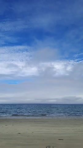 The video shows a wide shot of a beach and the ocean. The sky is blue with white clouds. The ocean is a deep blue color with gentle waves. The beach is sandy and appears to be empty. The video pans slowly across the scene, creating a serene and calming atmosphere.