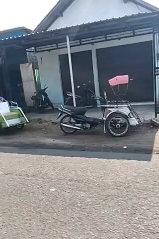 The video shows a street scene with several shops and a few people. In the foreground, a motorcycle with a sidecar is parked. A woman is seen on a motorcycle in the middle ground, wearing a maroon shirt and pink pants. Further down the street, a store with a blue and yellow sign is visible, as well as other small buildings. The street appears to be paved, and there is some greenery and debris along the sides. The overall impression is of a quiet, possibly rural or semi-urban, street with a few small businesses.