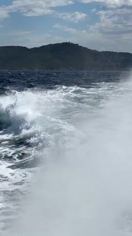 A video captures a choppy ocean with waves crashing in the foreground. In the distance, a green, hilly coastline is visible under a partly cloudy sky. The shot is taken from a moving vessel, indicated by the frothy wake trailing behind. No people or specific objects are discernible in the frame.
