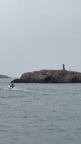 A speed boat with two people onboard is cruising through choppy gray water. The boat is moving from left to right across the frame. In the background, a rocky island with a small red lighthouse on top is visible. The sky is overcast and gray.