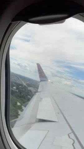 The video shows a view from an airplane window looking out at a wing and the landscape below. The wing is white with some grey markings and a red tip. The landscape is green with trees and some buildings, suggesting it is a rural or suburban area. The sky is cloudy. The video is short and appears to be a continuous shot.